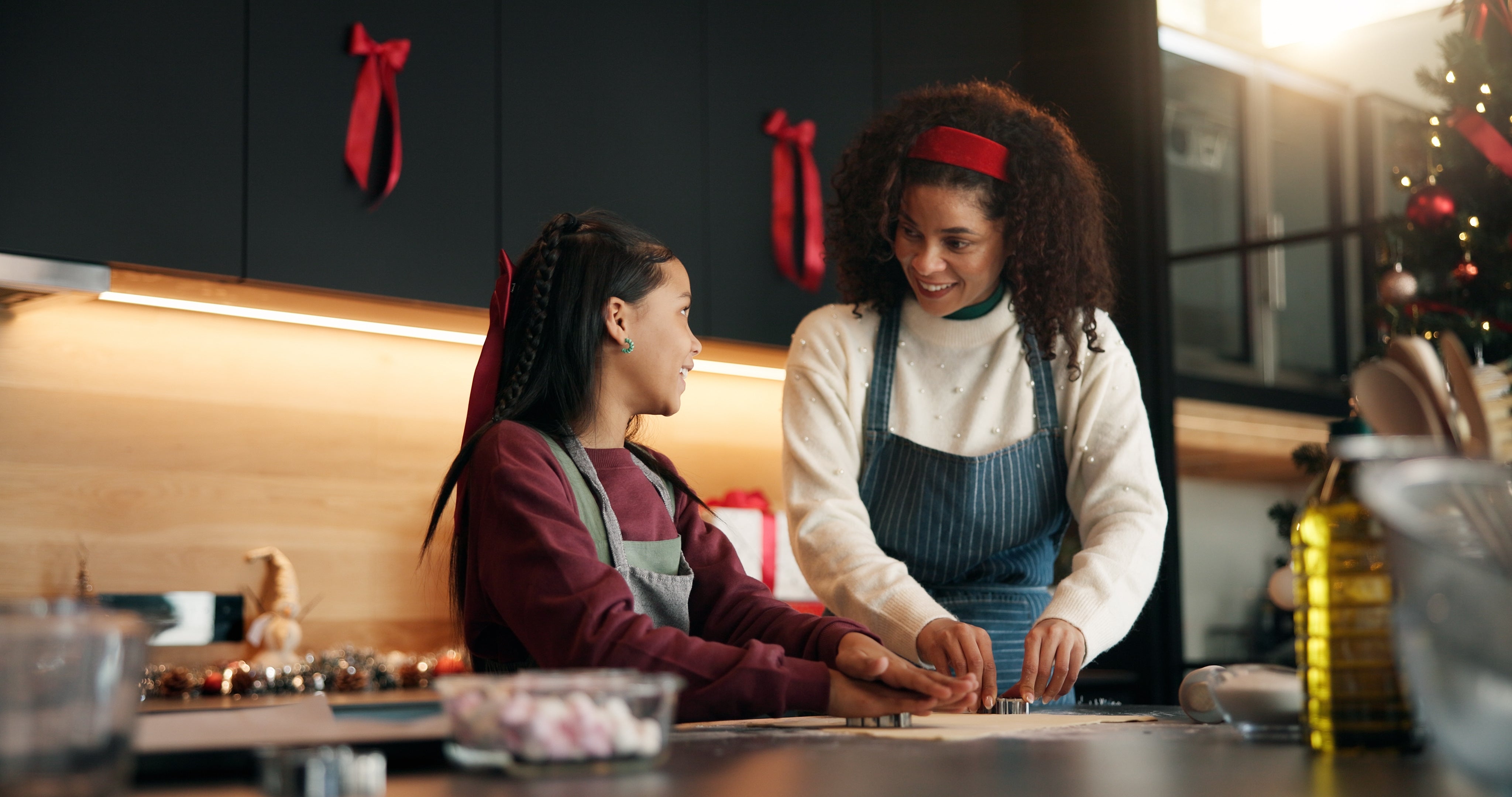 Two women in a kitchen preparing food, with festive decorations and under-cabinet lighting in the background.